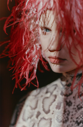 Portrait of a fair-skinned adult female with blue eyes, red feathered headwear, and an ornate snakeskin-patterned dress.