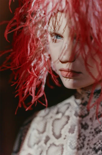 Portrait of a fair-skinned adult female with blue eyes, red feathered headwear, and an ornate snakeskin-patterned dress.