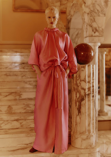 A fair-skinned person with a short blonde haircut poses on marble stairs, wearing a long, loose-fitting pink dress with pockets and brown shoes.