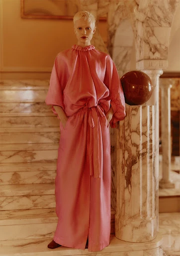 A fair-skinned person with a short blonde haircut poses on marble stairs, wearing a long, loose-fitting pink dress with pockets and brown shoes.