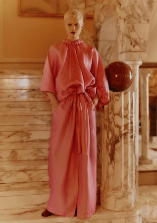 A fair-skinned person with a short blonde haircut poses on marble stairs, wearing a long, loose-fitting pink dress with pockets and brown shoes.