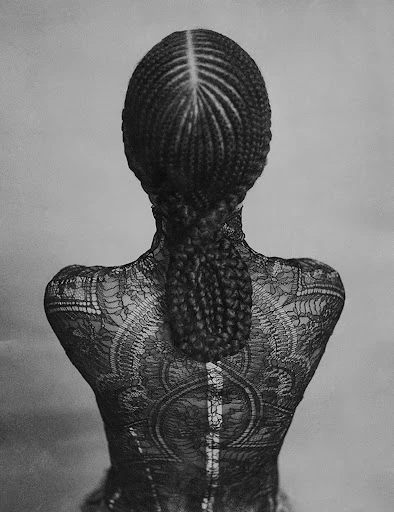 Black and white portrait from behind of an adult woman with cornrow braids, wearing a lace top, against a grey backdrop.