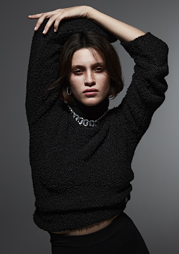 Model with short brown hair wearing a black textured turtleneck sweater and silver necklace against a grey backdrop