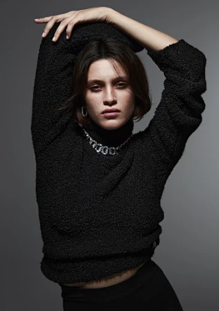 Model with short brown hair wearing a black textured turtleneck sweater and silver necklace against a grey backdrop