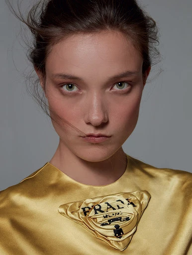 Close-up portrait of a young adult woman with fair skin and green eyes wearing a gold dress with a Prada logo patch on the chest.