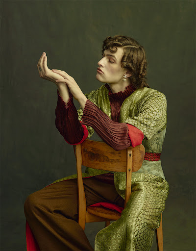 Fashion portrait of a young adult male sitting on a chair wearing a patterned robe, with hands up, in a studio with a neutral background.