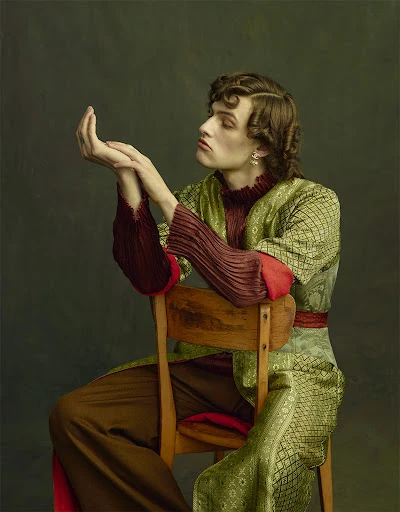 Fashion portrait of a young adult male sitting on a chair wearing a patterned robe, with hands up, in a studio with a neutral background.