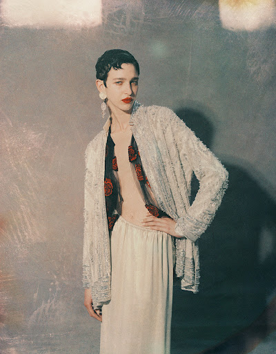 Fashion shot of a woman with short dark hair, wearing a glittering jacket and a long skirt, posing in a photo studio.