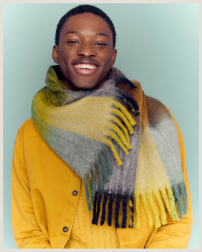 Portrait of a Black male smiling and wearing a yellow jacket and colorful fringed scarf against a light blue background in a studio.
