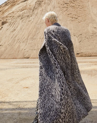A person wearing a spotted coat standing in a sandy environment with a large sand dune visible in the background on a sunny day.