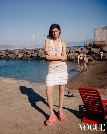 A young woman stands on a stone pier in a crochet top and skirt near the sea on a sunny day. The Vogue logo appears in the corner.