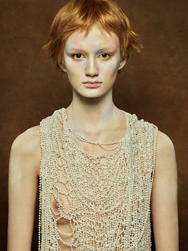 Eye-level portrait of an adult woman with short red hair, wearing heavy pearl jewelry, against a solid brown studio backdrop.