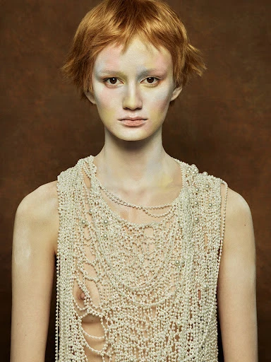 Eye-level portrait of an adult woman with short red hair, wearing heavy pearl jewelry, against a solid brown studio backdrop.
