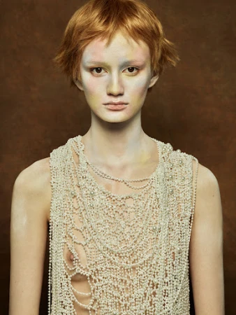 Eye-level portrait of an adult woman with short red hair, wearing heavy pearl jewelry, against a solid brown studio backdrop.