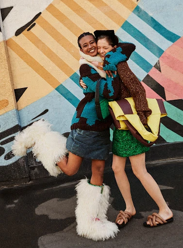 Two smiling adult females embrace outdoors; one wears a blue-and-brown patterned sweater, a denim skirt, and white furry boots; the other a brown sweater and green skirt.