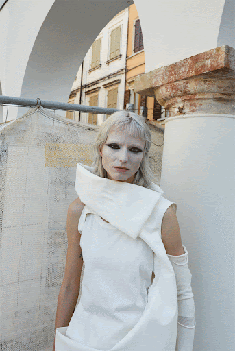 Albino fashion model with dramatic eye makeup poses in a white ensemble against an outdoor urban backdrop with architectural elements.