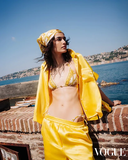 Fashion shot of a woman in a patterned bikini and yellow silk shirt and pants, posing outdoors with a coastal background and blue sky.