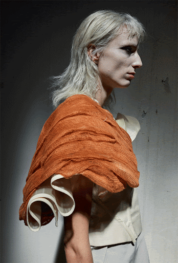 A person with light hair wears an orange textured cape with a white garment in an indoor environment against a grey wall.