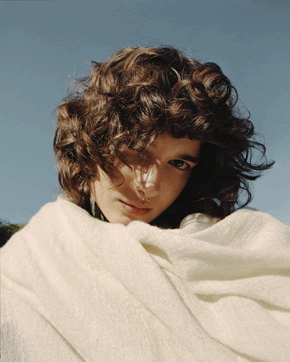 Close-up portrait of a young adult woman with curly brown hair wrapped in a textured white blanket under clear blue sky outdoors.