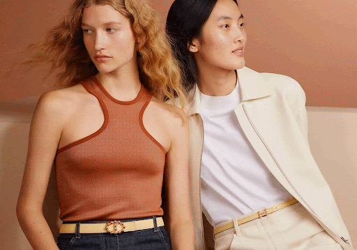 Two fashion models pose indoors with a light-brown backdrop. One woman wears a brown tank top and jeans; the other wears a white jacket and pants.