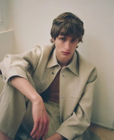 Young man in beige collared jacket and trousers sitting indoors by a plain wall