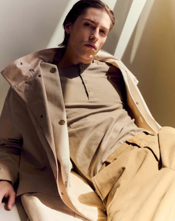 A young man wearing a neutral henley shirt and tan coat is captured in warm sunlight with harsh shadow lines.