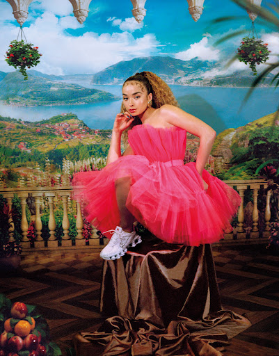 Adult female with curly brown hair, sitting on a draped pedestal, wearing a pink dress and white sneakers, against an ornate background.