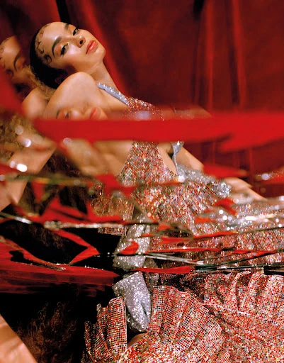 A young woman with dark hair and beaded dress poses against a red curtain, with reflections adding depth to a fashion-forward image.