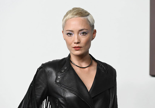 Close-up portrait of a woman with short blonde hair wearing a black leather jacket and necklace against a neutral backdrop. 