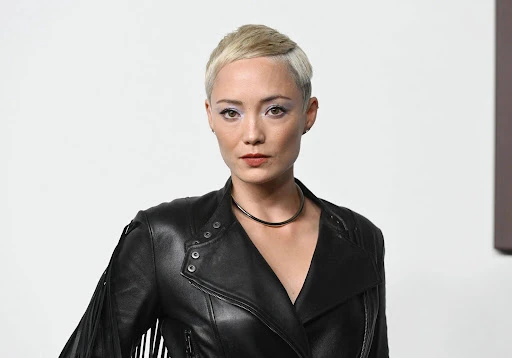 Close-up portrait of a woman with short blonde hair wearing a black leather jacket and necklace against a neutral backdrop. 