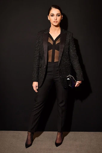 Naomi Scott poses in an all-black pantsuit with a sheer top against a dark background, holding a clutch, at a celebrity event.