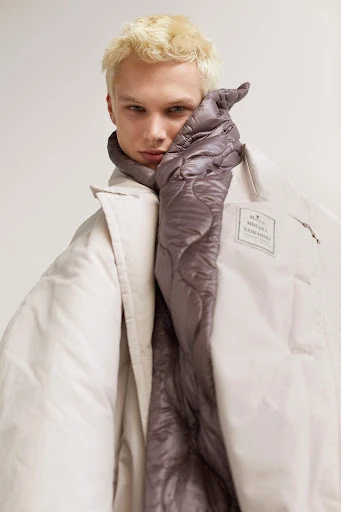 Fair-skinned male model with blond hair wears an oversized, pale gray puffer jacket with a quilted gray lining, studio lighting.