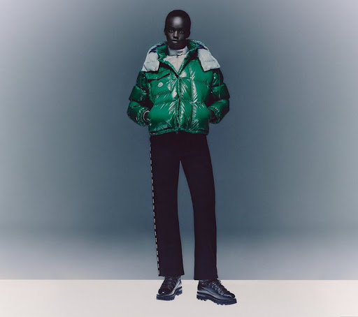A dark-skinned model presents a shiny green puffer jacket with a light grey hood, paired with dark pants and chunky shoes in a studio setting.