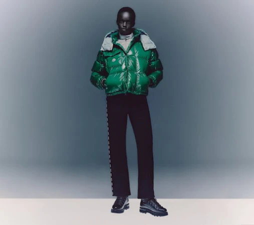 A dark-skinned model presents a shiny green puffer jacket with a light grey hood, paired with dark pants and chunky shoes in a studio setting.