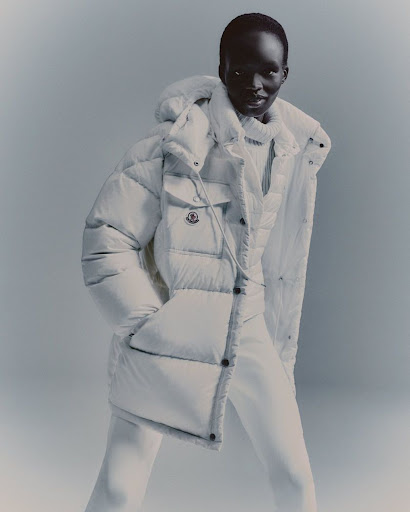 Studio shot of a dark-skinned adult female fashion model posing in a puffy white hooded coat with white pants against a white backdrop.