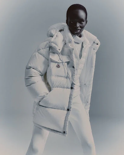 Studio shot of a dark-skinned adult female fashion model posing in a puffy white hooded coat with white pants against a white backdrop.