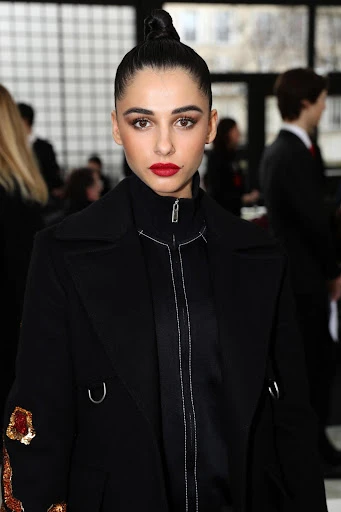 Close-up portrait of a woman with her hair in a bun, wearing a black coat, posing indoors at a fashion show or event. Red lipstick.