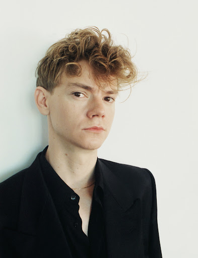 Portrait of a man with curly light brown hair wearing a black button-down shirt against a plain white wall