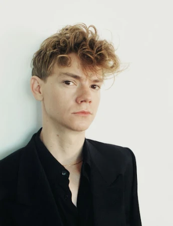 Portrait of a man with curly light brown hair wearing a black button-down shirt against a plain white wall