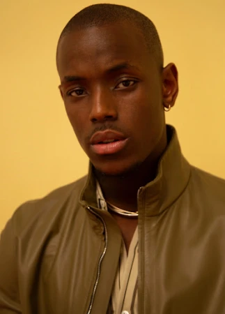 Close-up portrait of a man wearing a high-collared olive green jacket against a warm yellow background.