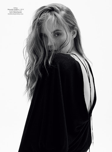 Black-and-white portrait of a young adult woman with long blonde hair, looking over her shoulder, wearing a black top, against a white background.