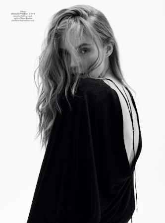 Black-and-white portrait of a young adult woman with long blonde hair, looking over her shoulder, wearing a black top, against a white background.
