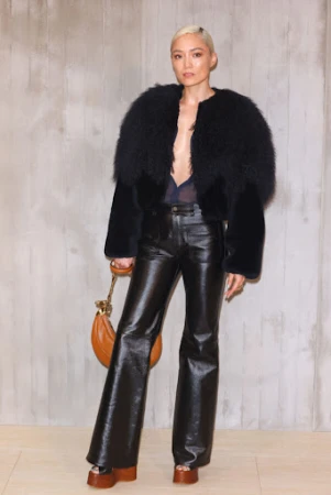 Fashionable woman in a fur-trimmed jacket, shiny black flared pants, and platform heels, poses against a textured neutral wall with a purse.