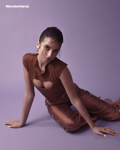 Attractive young adult female fashion model poses in cut-out top and sequined pants on purple backdrop looking at the camera with neutral expression.