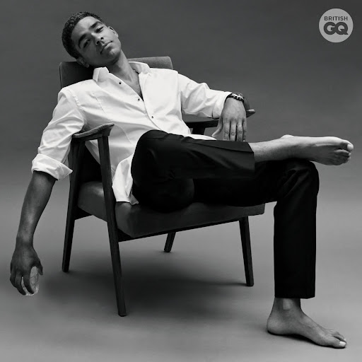 A man in a white shirt and black pants reclines in a chair. The photo is black and white with the British GQ logo in the corner.