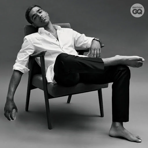 A man in a white shirt and black pants reclines in a chair. The photo is black and white with the British GQ logo in the corner.