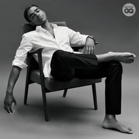 A man in a white shirt and black pants reclines in a chair. The photo is black and white with the British GQ logo in the corner.