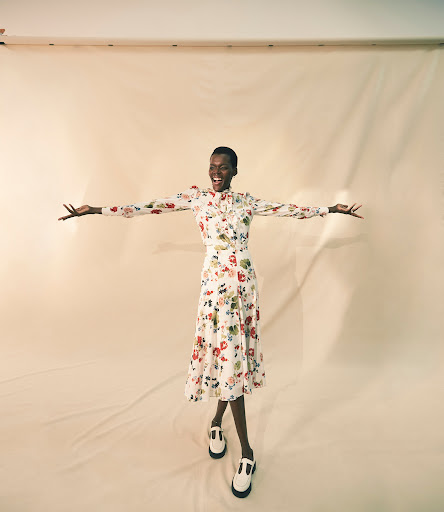 A joyful Black female model in a floral dress with arms outstretched, wears T-strap shoes, against a light backdrop, studio lighting.