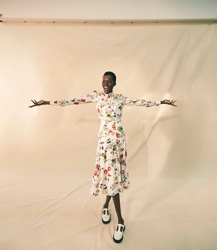 A joyful Black female model in a floral dress with arms outstretched, wears T-strap shoes, against a light backdrop, studio lighting.