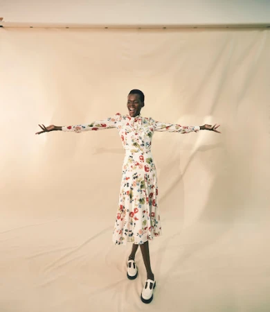 A joyful Black female model in a floral dress with arms outstretched, wears T-strap shoes, against a light backdrop, studio lighting.